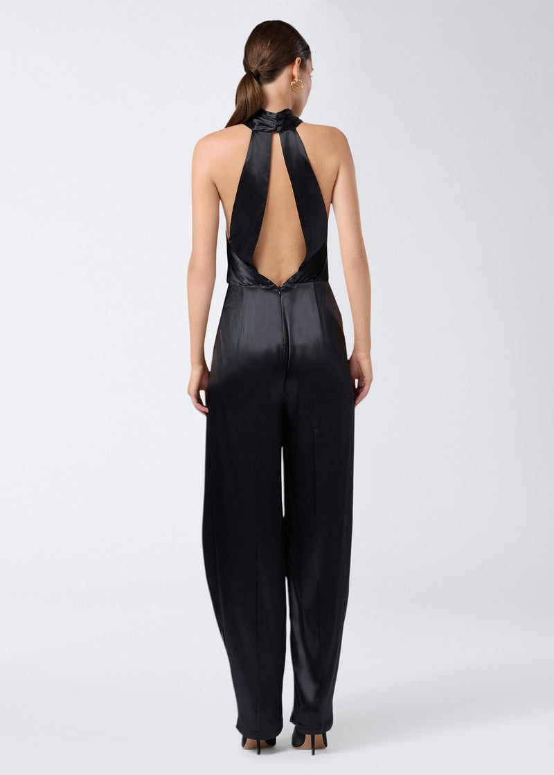 
                          Rear view of the Zeyne Jumpsuit, featuring black shiny satin fabric, cross-over halterneck straps, open back, and tapered balloon pants with pleats.
                        