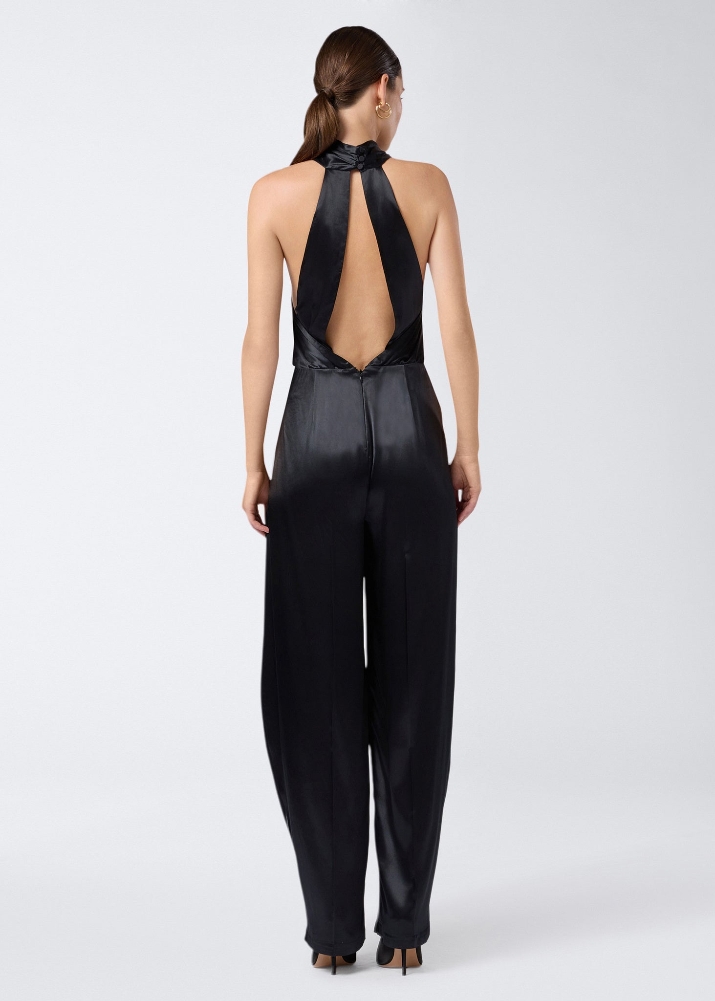 Rear view of the Zeyne Jumpsuit, featuring black shiny satin fabric, cross-over halterneck straps, open back, and tapered balloon pants with pleats.