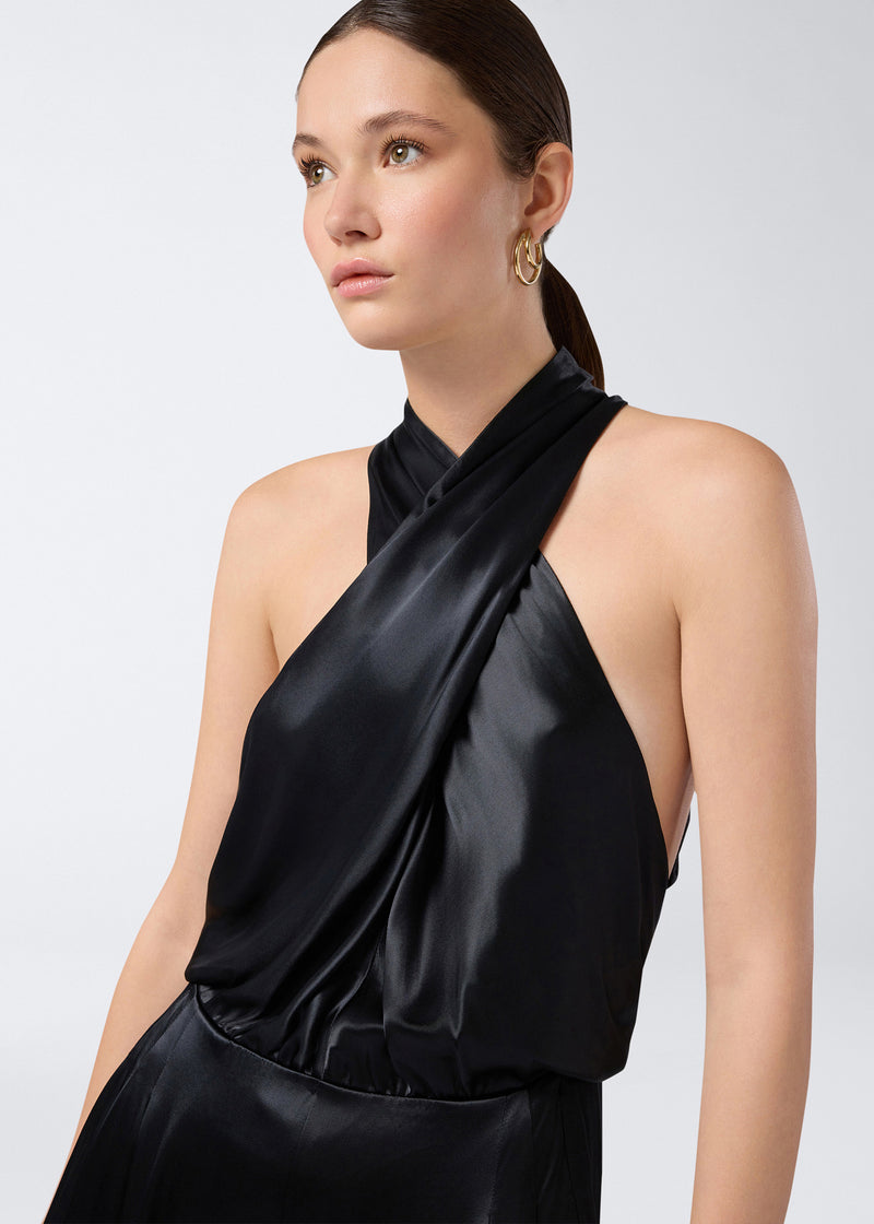 
                          Frontal view showing a model wearing the Zeyne Jumpsuit in shiny black satin. The jumpsuit features cross-over halterneck straps and a elegant sleeveless design.
                        