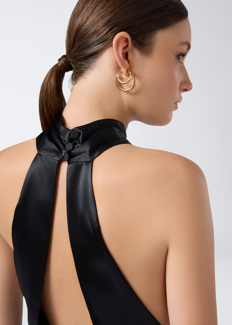 
                          Detail view of the Zeyne Jumpsuit, focusing on the black shiny satin halterneck straps fastened by buttons at the back of a woman&#39;s neck.
                        