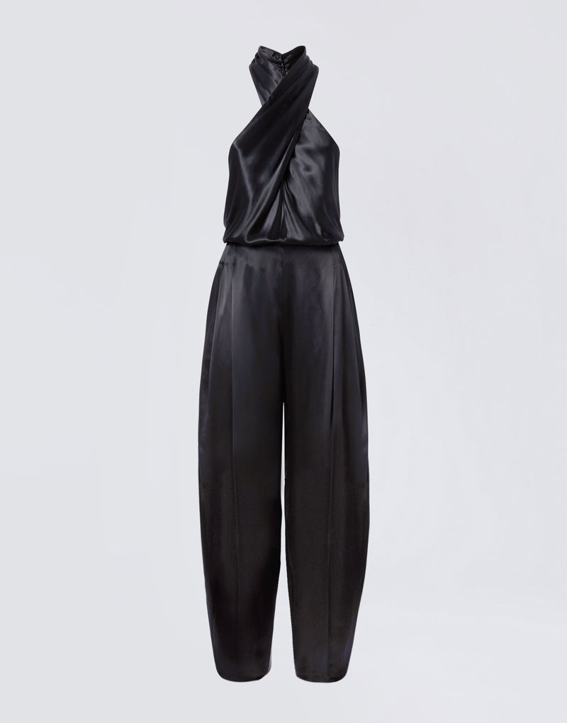
                          Frontal view of the Zeyne Jumpsuit, showing black shiny satin fabric, cross-over halterneck straps, and tapered balloon pants with front pleats.
                        