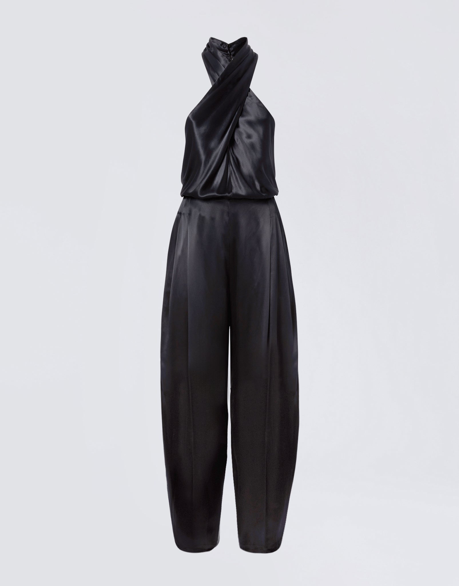Frontal view of the Zeyne Jumpsuit, showing black shiny satin fabric, cross-over halterneck straps, and tapered balloon pants with front pleats.