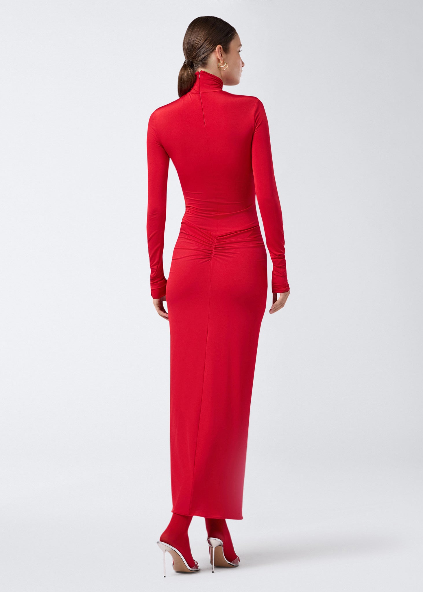 Rear view: Model stands wearing the Warwick Maxi Dress in red lightweight jersey, featuring a turtleneck, long sleeves, and waist ruching with a mermaid-effect skirt.