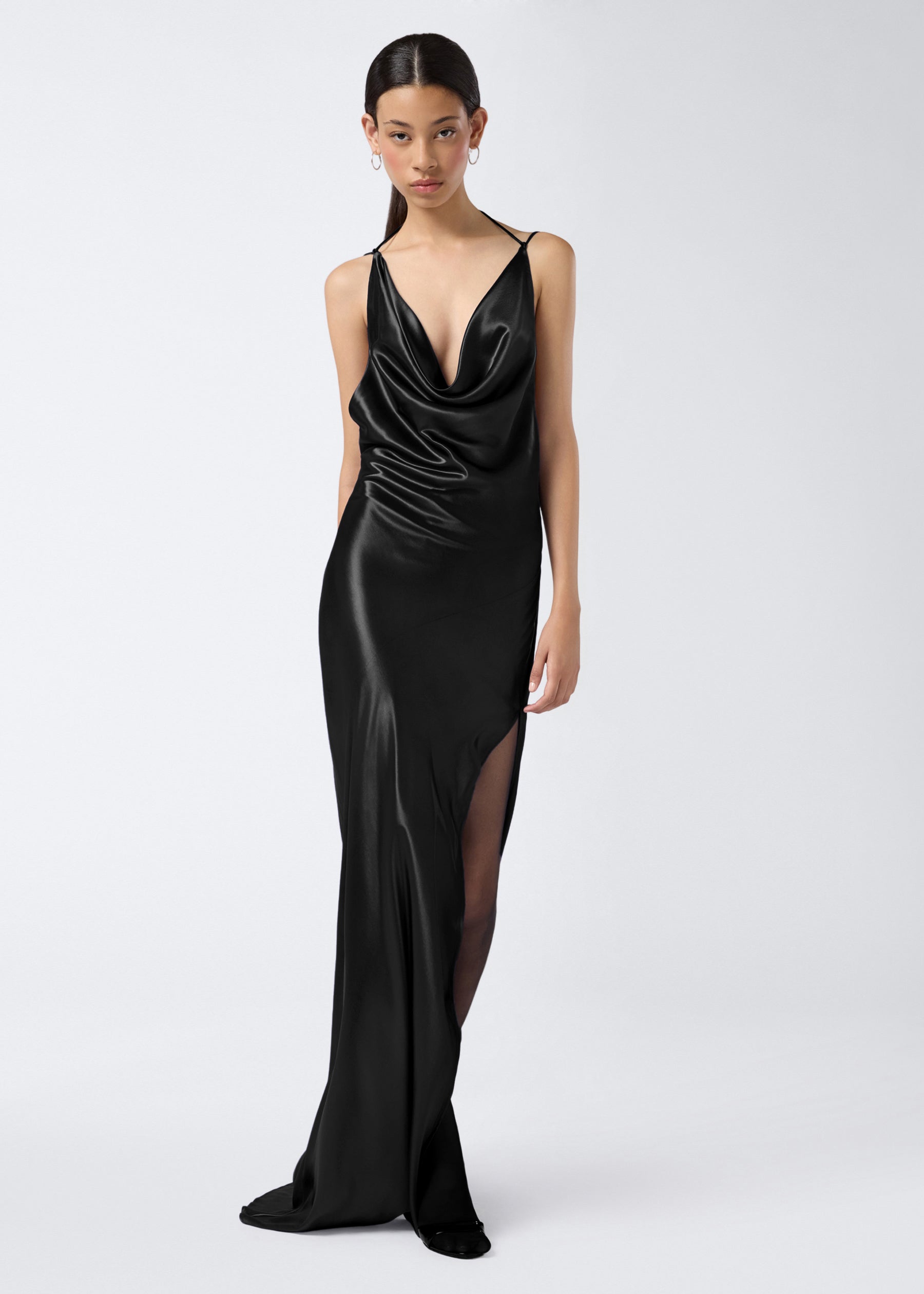 Frontal view of a model wearing the Vera Maxi Dress, a floor-length black shiny satin gown with a plunging cowl neck, adjustable straps, and side slit.