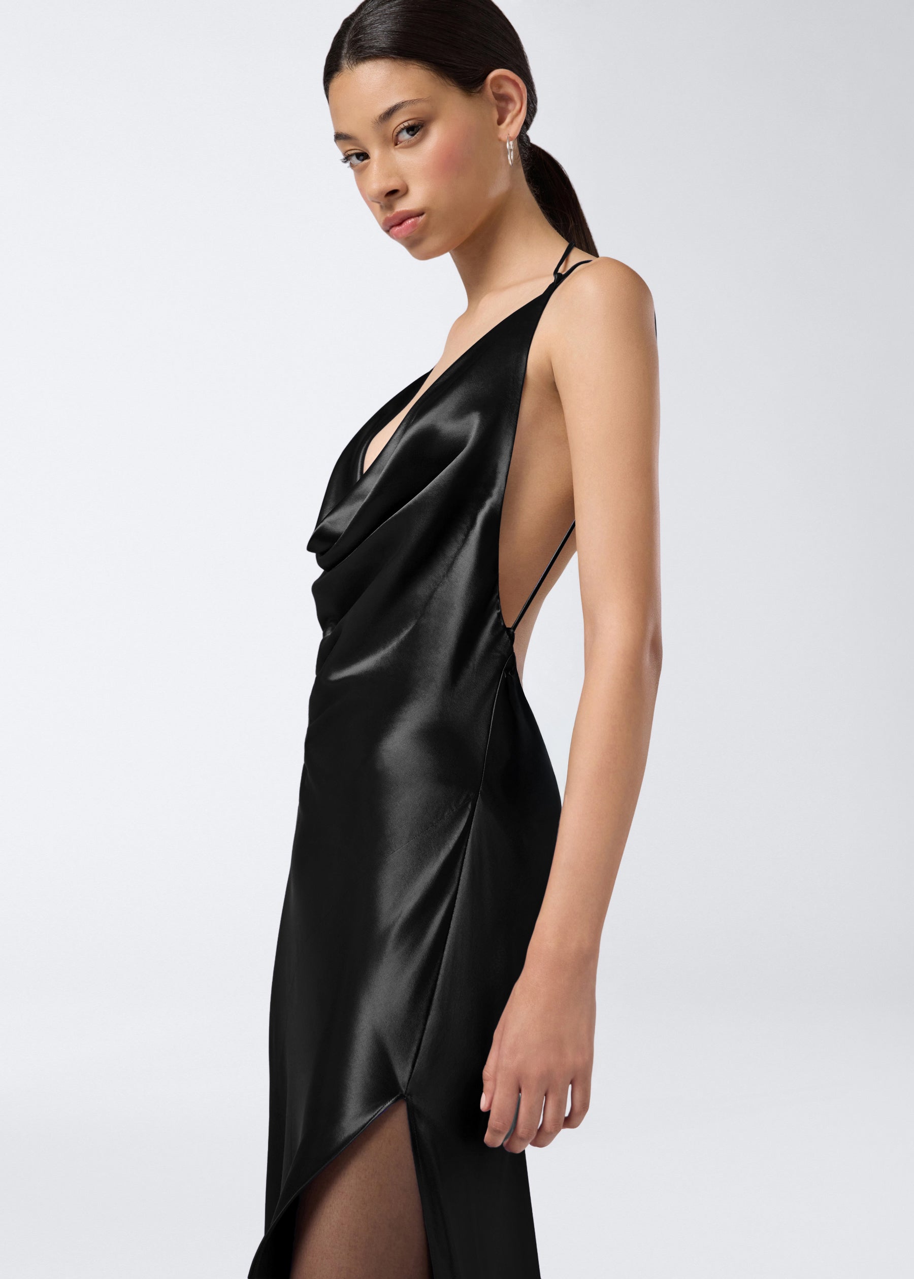 Side view of a woman wearing the Vera Maxi Dress in black shiny satin, featuring a plunging cowl neck, adjustable straps, and a floor-length cut.