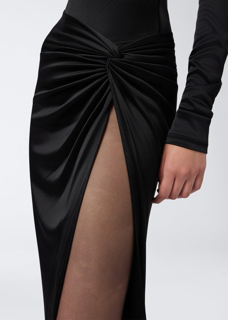 
                          Detail view of the Zohra Maxi Skirt showing black shiny jersey fabric with dramatic draping, a side knot detail, and a high slit revealing the leg.
                        