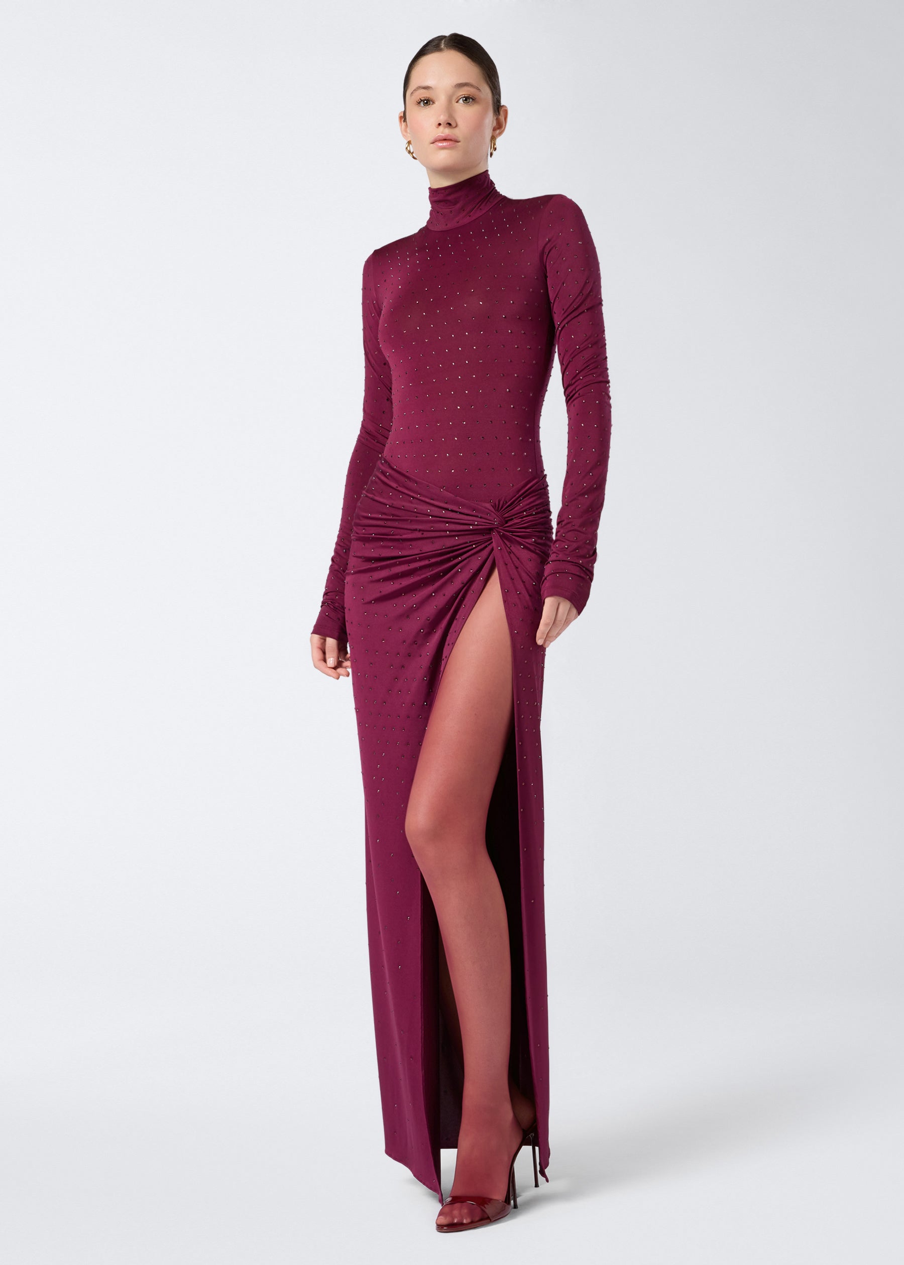 Frontal view of a woman wearing the Zohra Crystal Maxi Skirt in cabernet, featuring all-over crystals, draping with a knot detail at the side, and high slit.