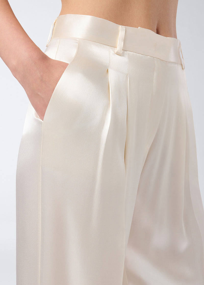 
                          Detail view of Nathalie Pants showing ivory shiny satin fabric, pleated front, high-rise waist, belt loops, and side slit pocket with hand inside.
                        