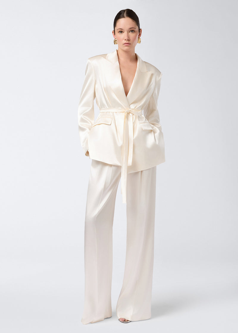 
                          Frontal view of Nathalie Pants in ivory shiny satin, featuring wide hems, front pleats, high-rise waist, and side slit pockets, worn by a model.
                        