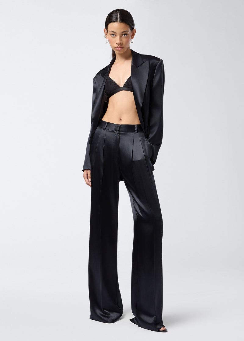 
                          Frontal view of a woman wearing the Nathalie Pants, featuring black shiny satin wide-hem design with front pleats, belt loops, and side slit pockets.
                        