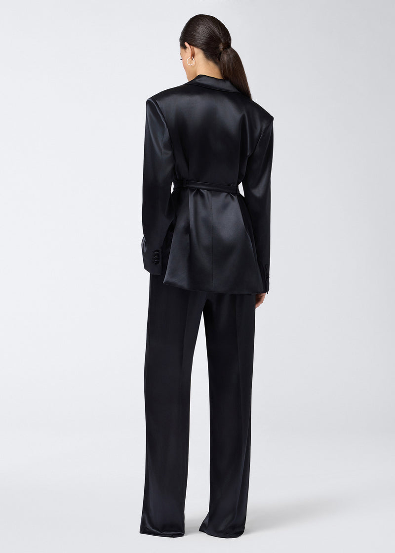 
                          Rear view of a woman wearing Nathalie Pants, featuring wide hems in shiny black satin, paired with a matching long blazer, both draping elegantly.
                        