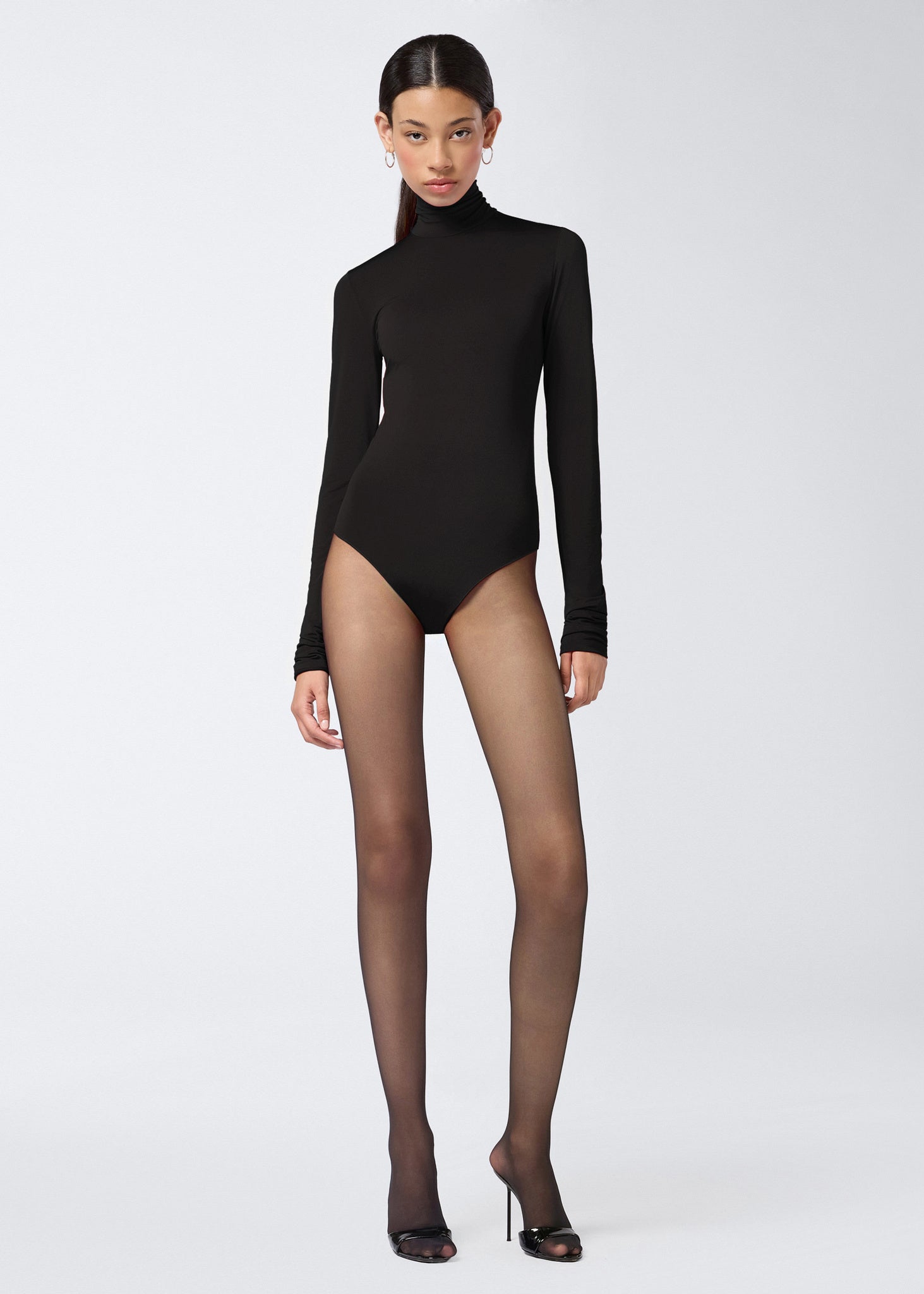 Frontal view of a model wearing the Ziggy Bodysuit, a black lightweight jersey piece with long sleeves and a turtleneck, styled with sheer black tights and heels.