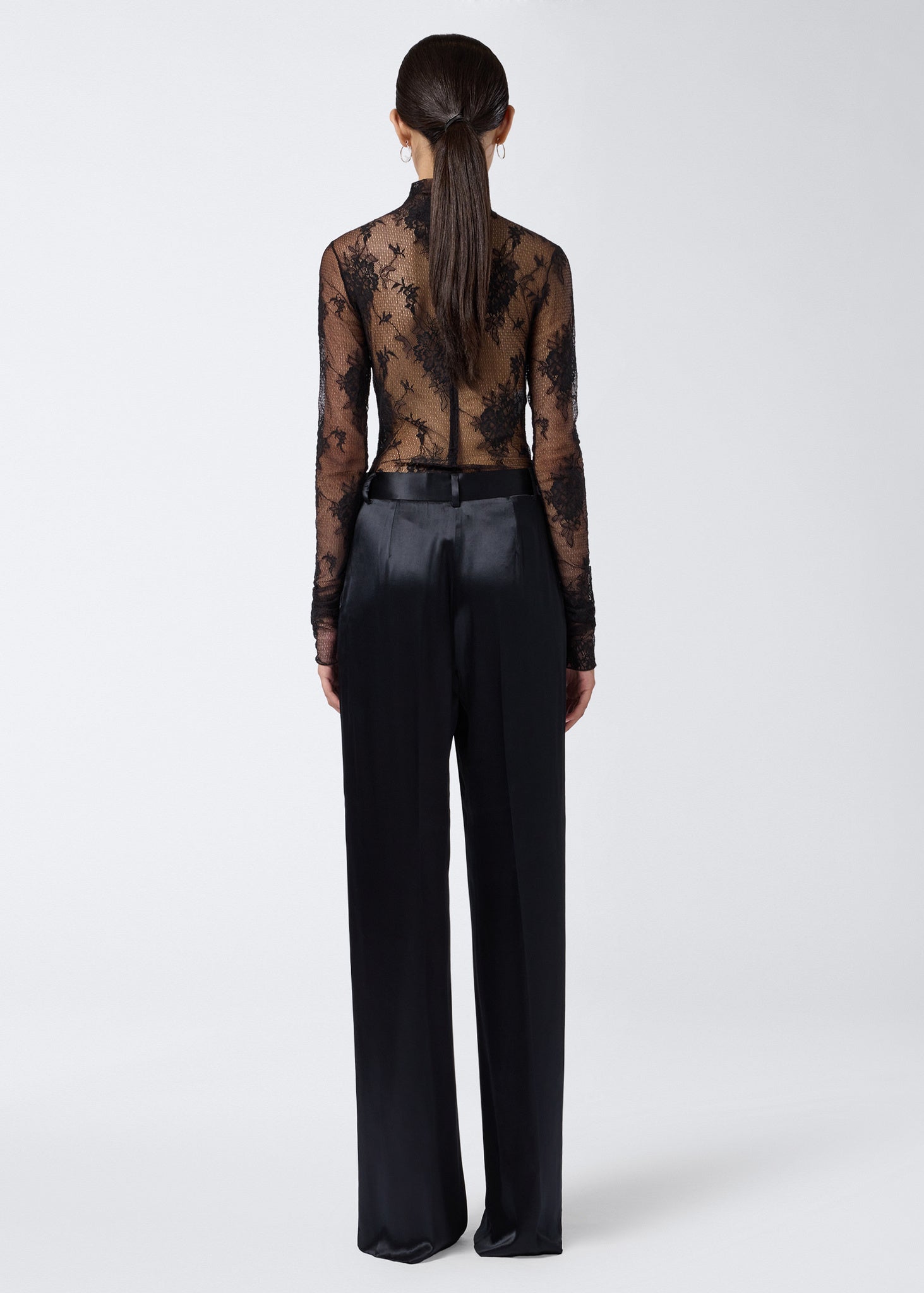 Rear view shows model wearing Ziggy Bodysuit, a sheer black lace, long-sleeve, turtleneck bodysuit, zipped at the back, paired with black satin pants.