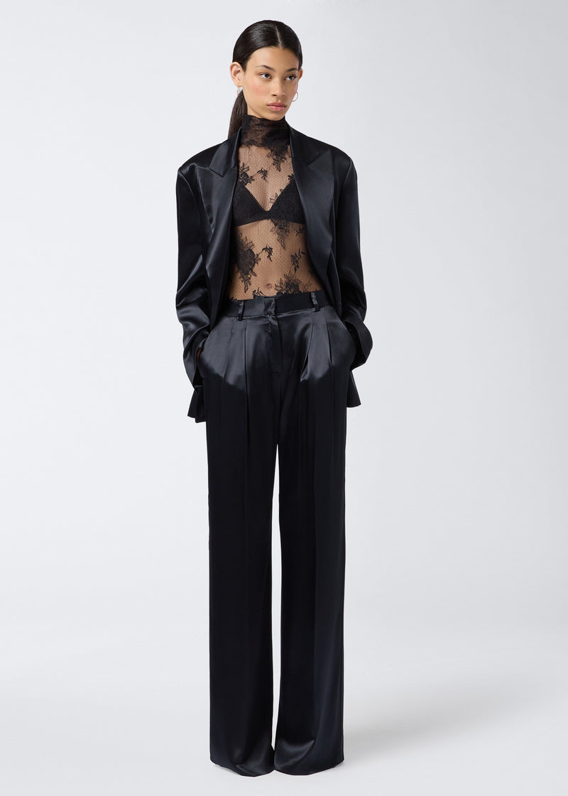 
                          Frontal view of a woman wearing the New Guenda Bra, made from black lace, visible under a sheer lace top, styled with black satin trousers and jacket.
                        