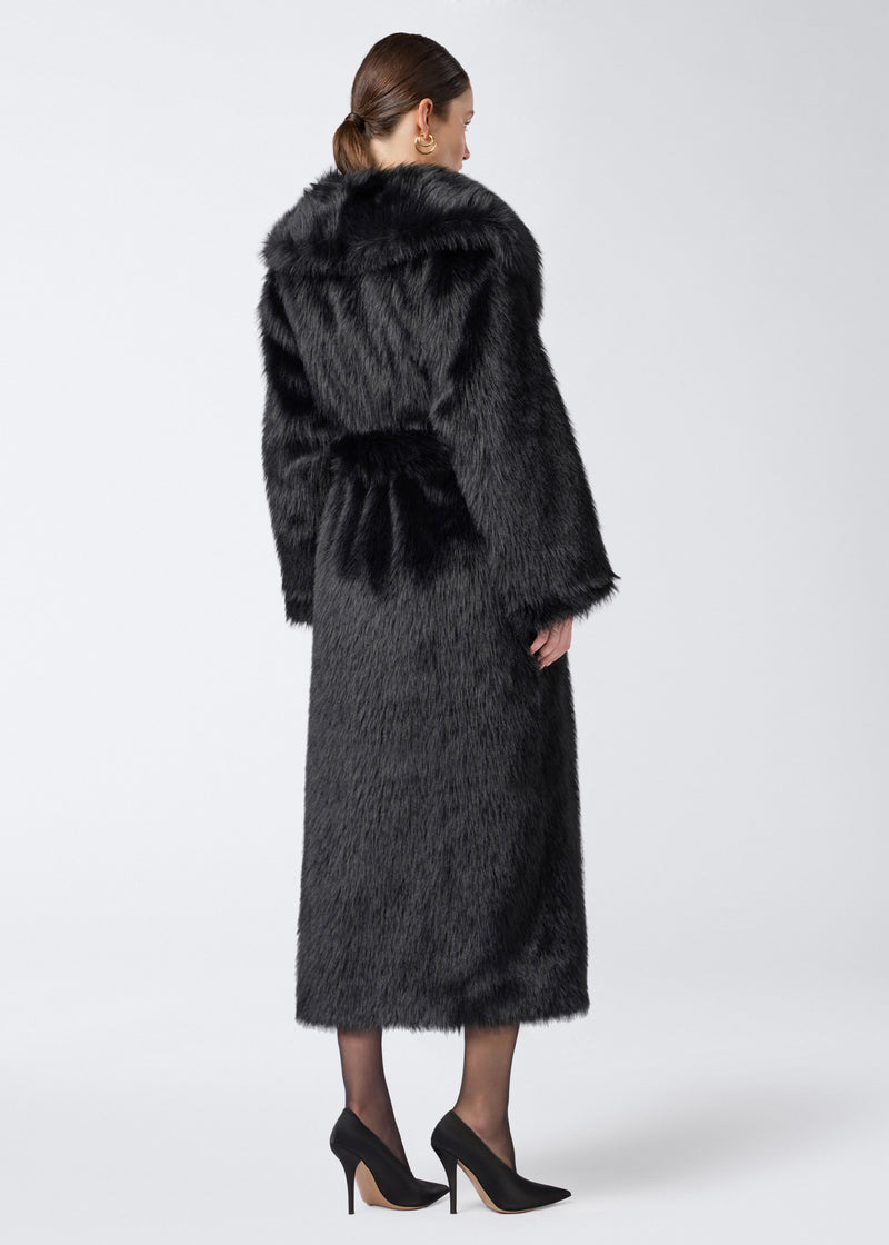 
                          Rear view: Model wearing the Winter Maxi Coat in black faux fur. The coat extends to the calves, features a large shawl lapel collar, long sleeves, and a matching waist belt.
                        