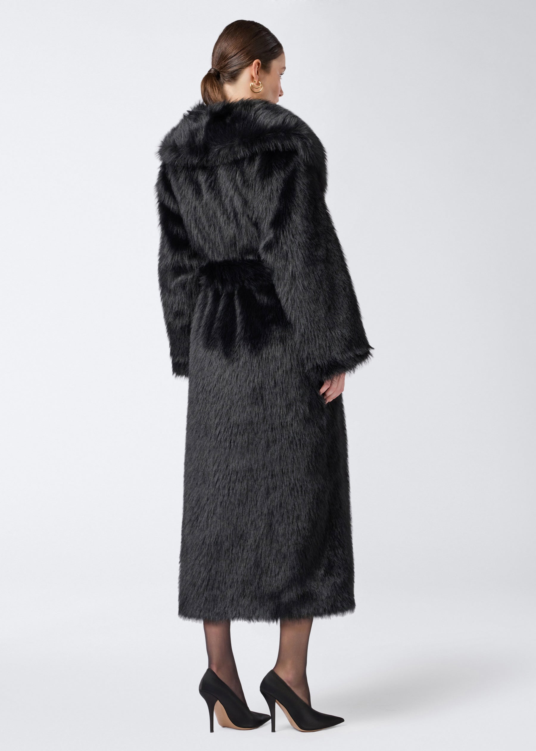 Rear view: Model wearing the Winter Maxi Coat in black faux fur. The coat extends to the calves, features a large shawl lapel collar, long sleeves, and a matching waist belt.