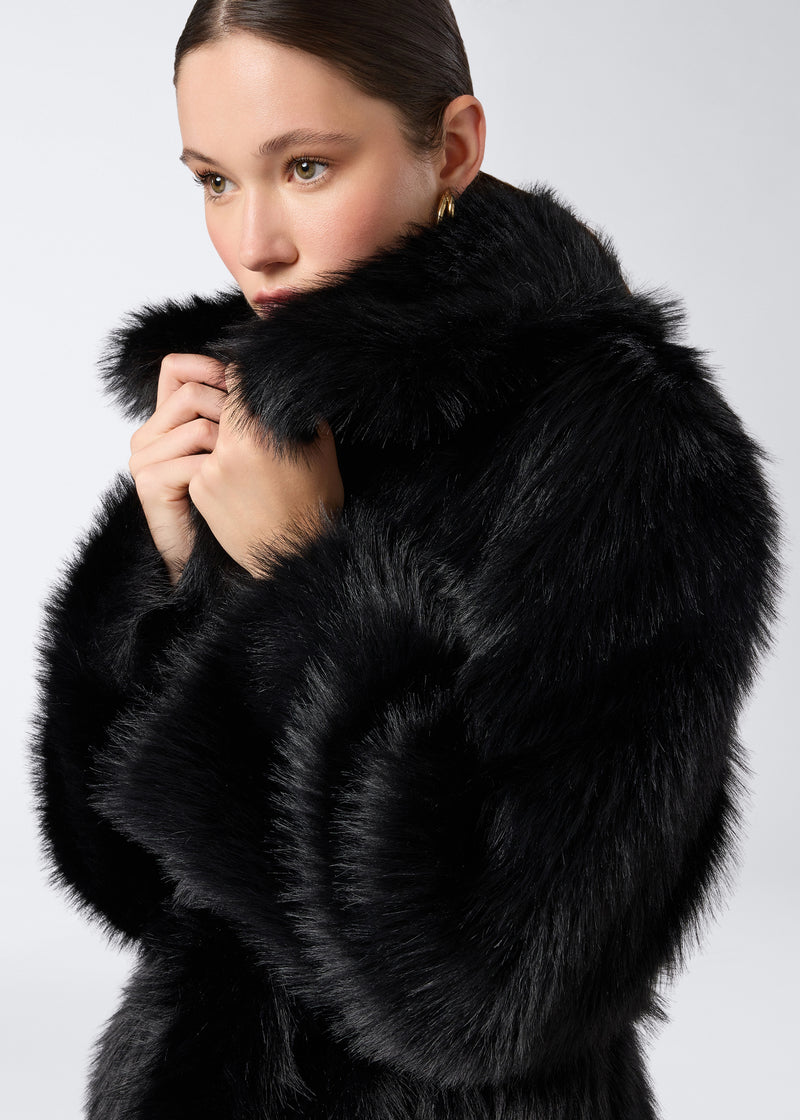 
                          Detail view. Woman holding up collar of Winter Maxi Coat in black faux fur, showing the plush texture, large shawl lapel collar, and long sleeves.
                        