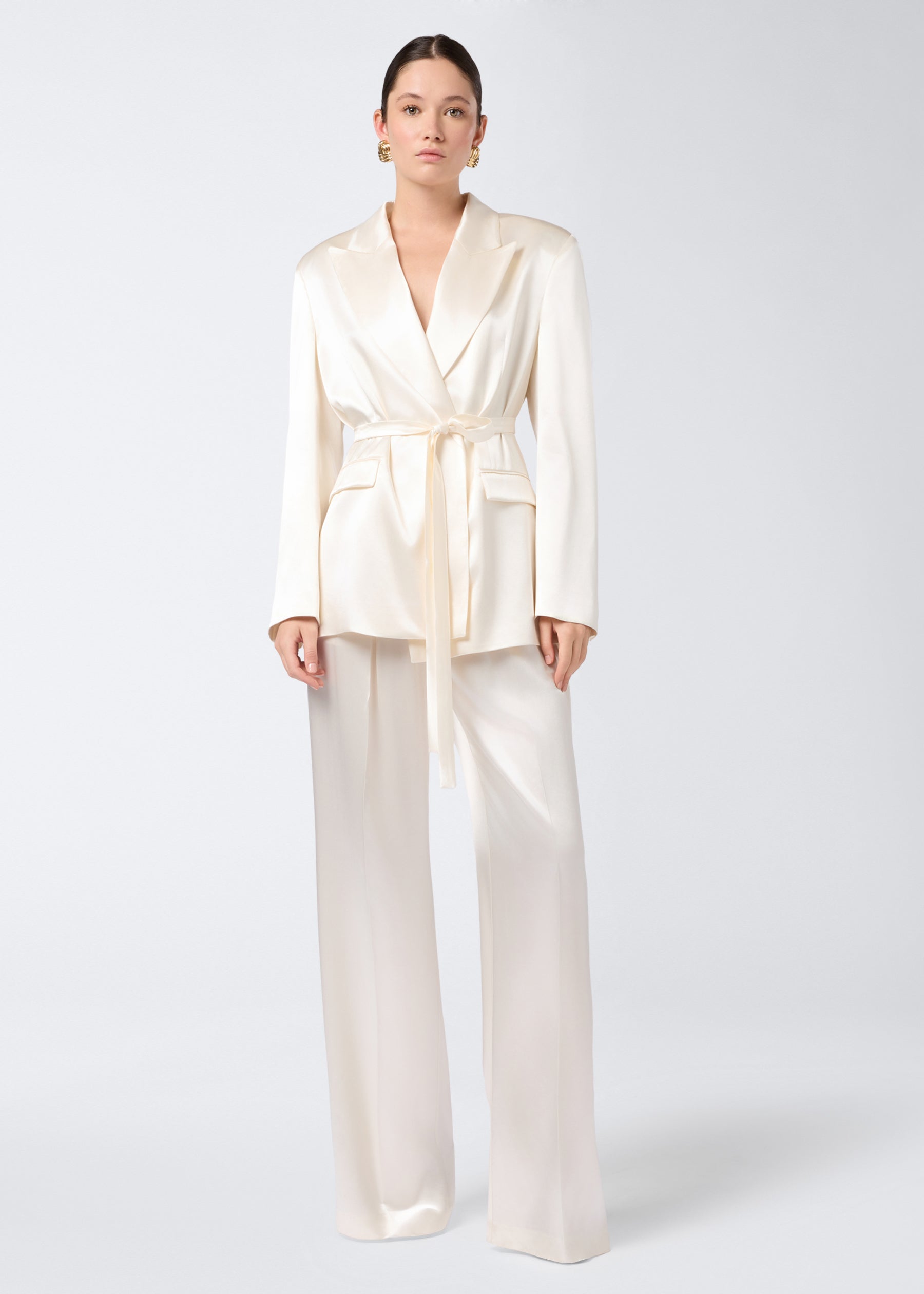 Frontal view showing model wearing Wilder Blazer in ivory shiny satin, with peaked lapel, belted waist, padded shoulders, two flap pockets, and buttoned cuffs.