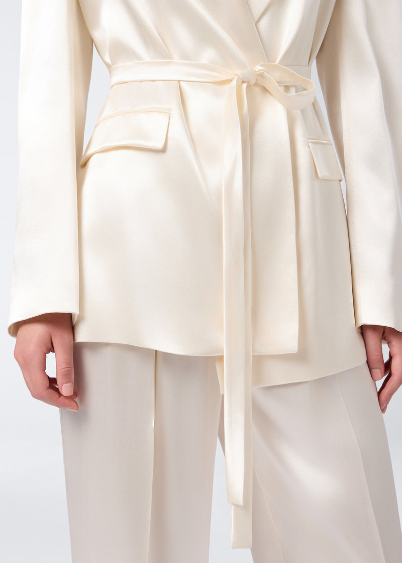 
                          Frontal view showing the waist-down detail of the Wilder Blazer in ivory shiny satin, featuring a tied belt, peaked lapel, flap pockets, and buttoned cuffs.
                        