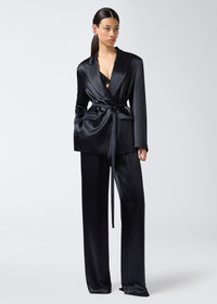 Frontal view of a woman wearing the Wilder Blazer in black shiny satin, featuring a matching belt at the waist, peaked lapel collar, flap pockets, long sleeves, and buttoned cuffs.