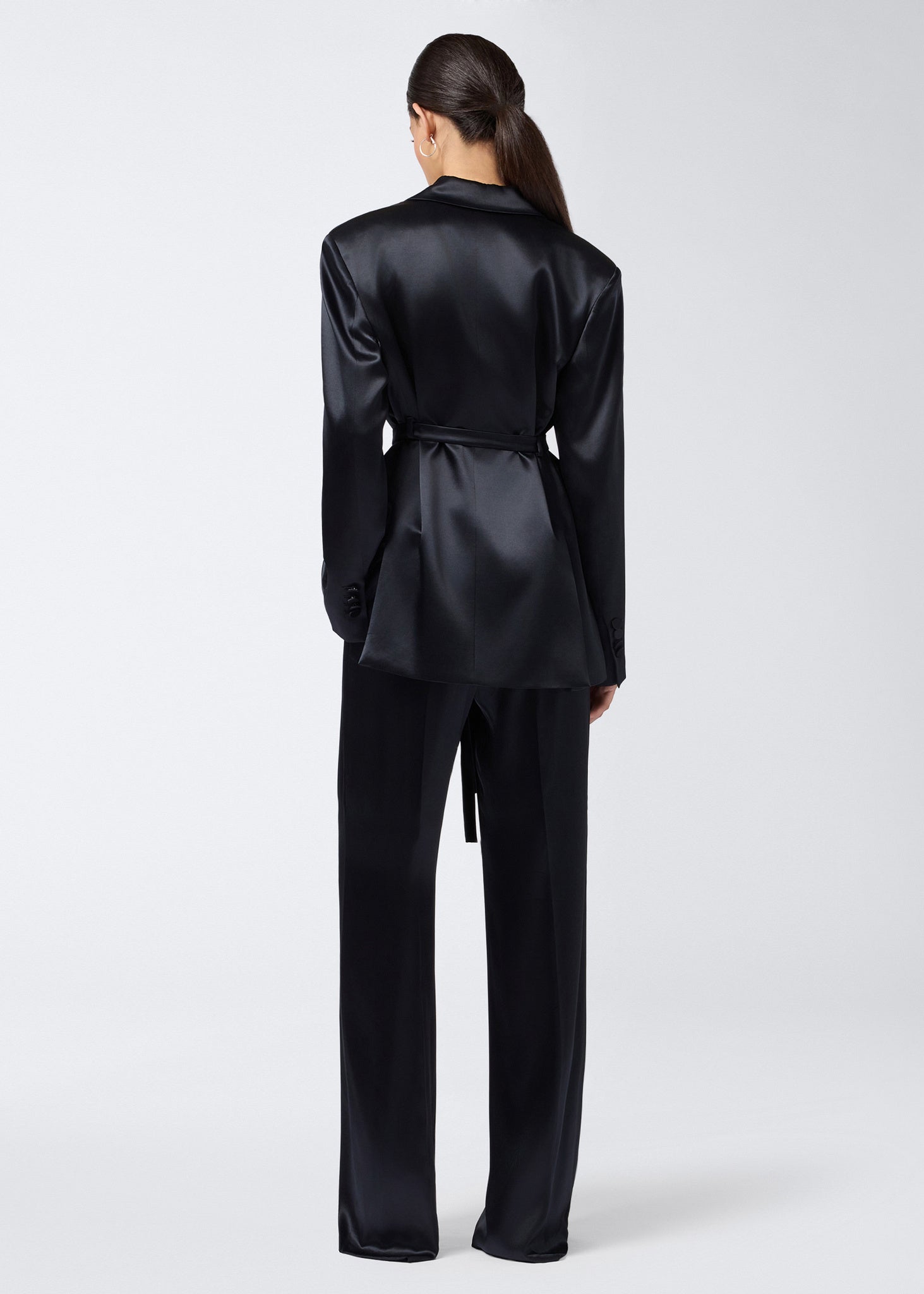 Rear view of a woman wearing the Wilder Blazer in black shiny satin, featuring a waist belt, long sleeves, and tailored structure with padded shoulders.