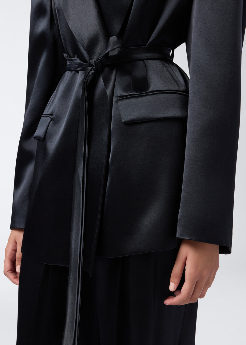 
                          Detail view of the Wilder Blazer, showcasing its black shiny satin fabric, matching waist belt, two waist flap pockets, and long sleeves on a woman.
                        