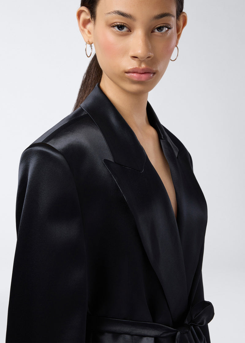 
                          Frontal view shows model wearing the Wilder Blazer in black shiny satin with a peaked lapel collar, padded shoulders, and matching waist belt.
                        