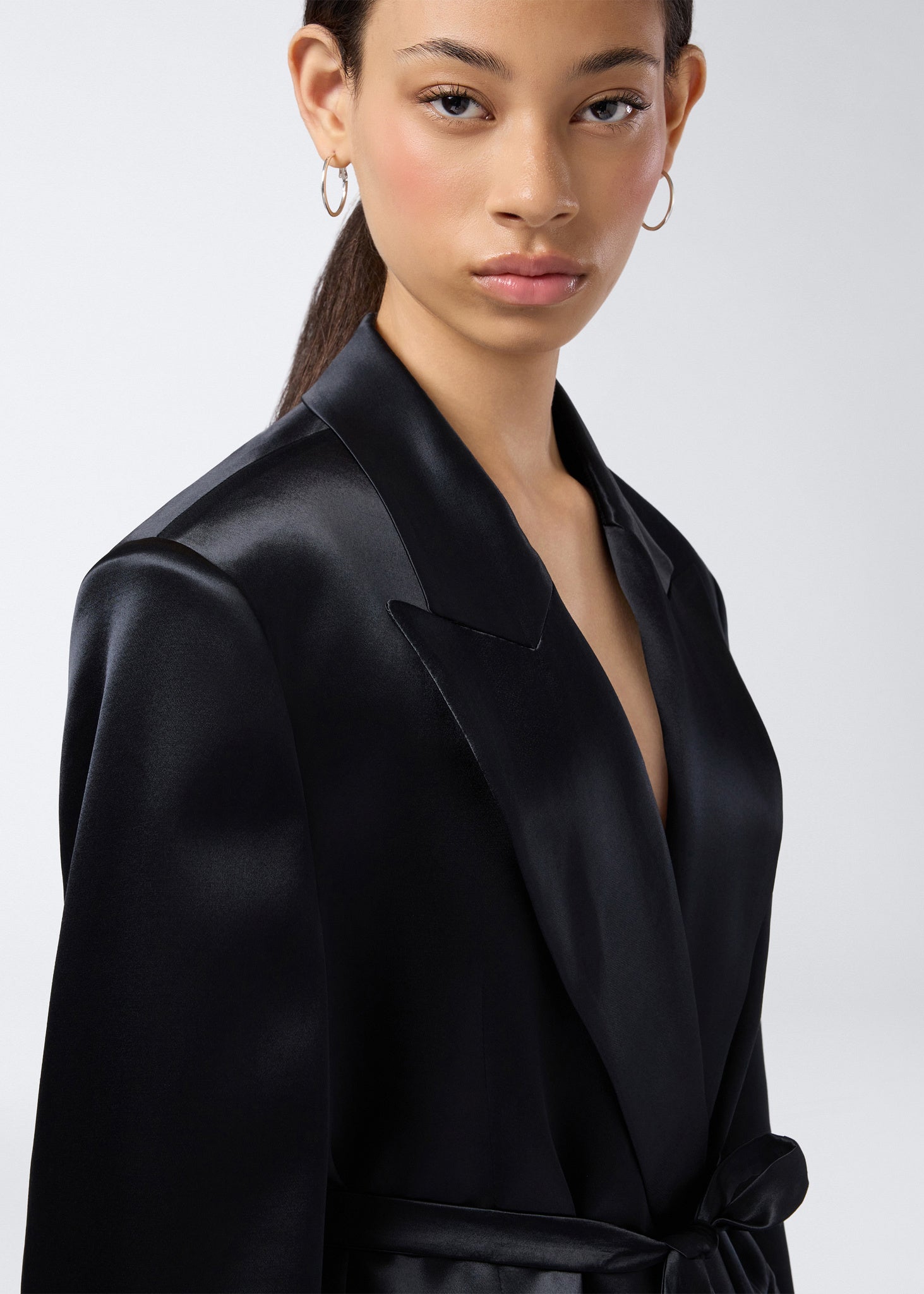 Frontal view shows model wearing the Wilder Blazer in black shiny satin with a peaked lapel collar, padded shoulders, and matching waist belt.