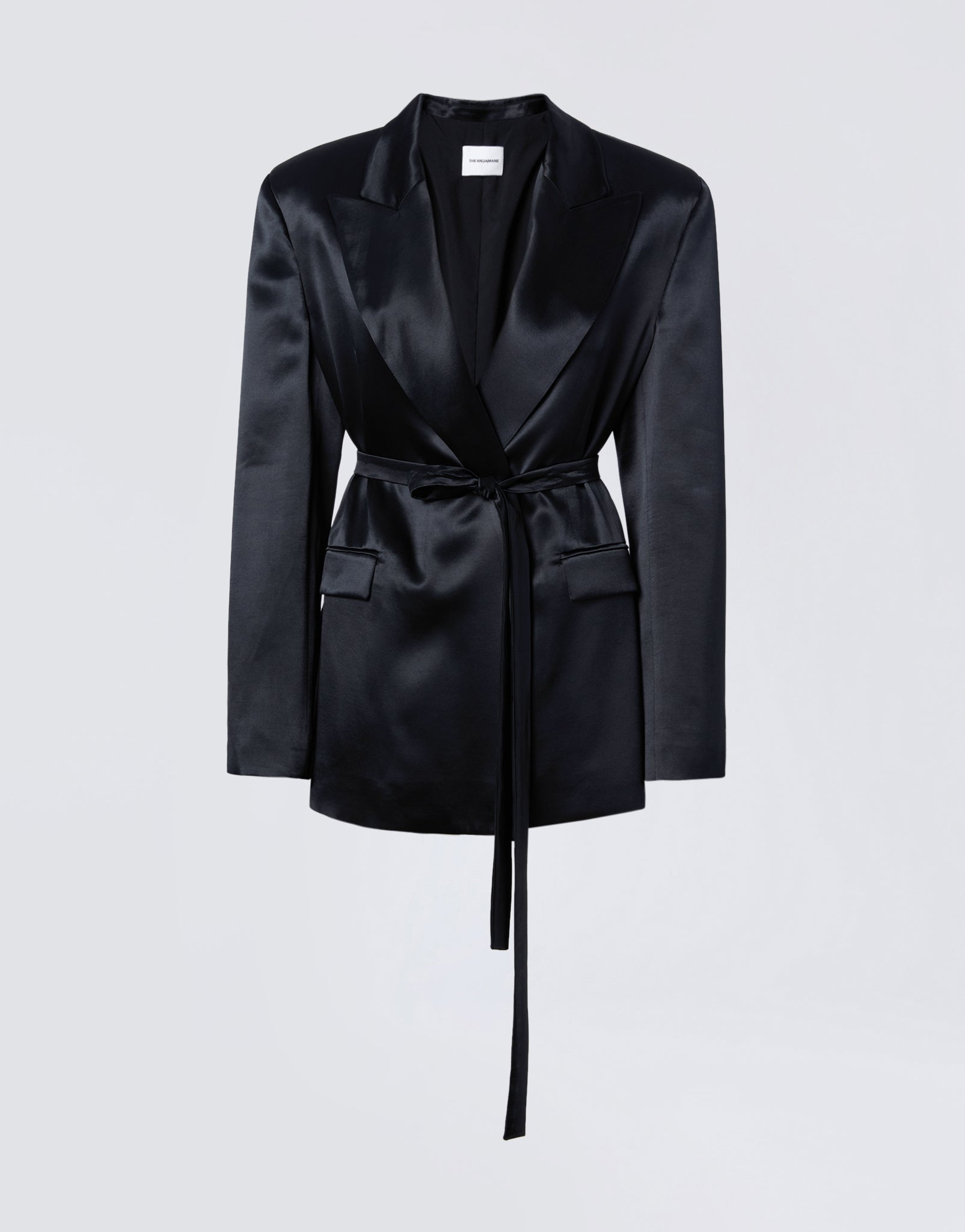 Frontal view of the Wilder Blazer, a black shiny satin women’s blazer with peaked lapel collar, padded shoulders, matching waist belt, two flap pockets, and long sleeves with buttoned cuffs.