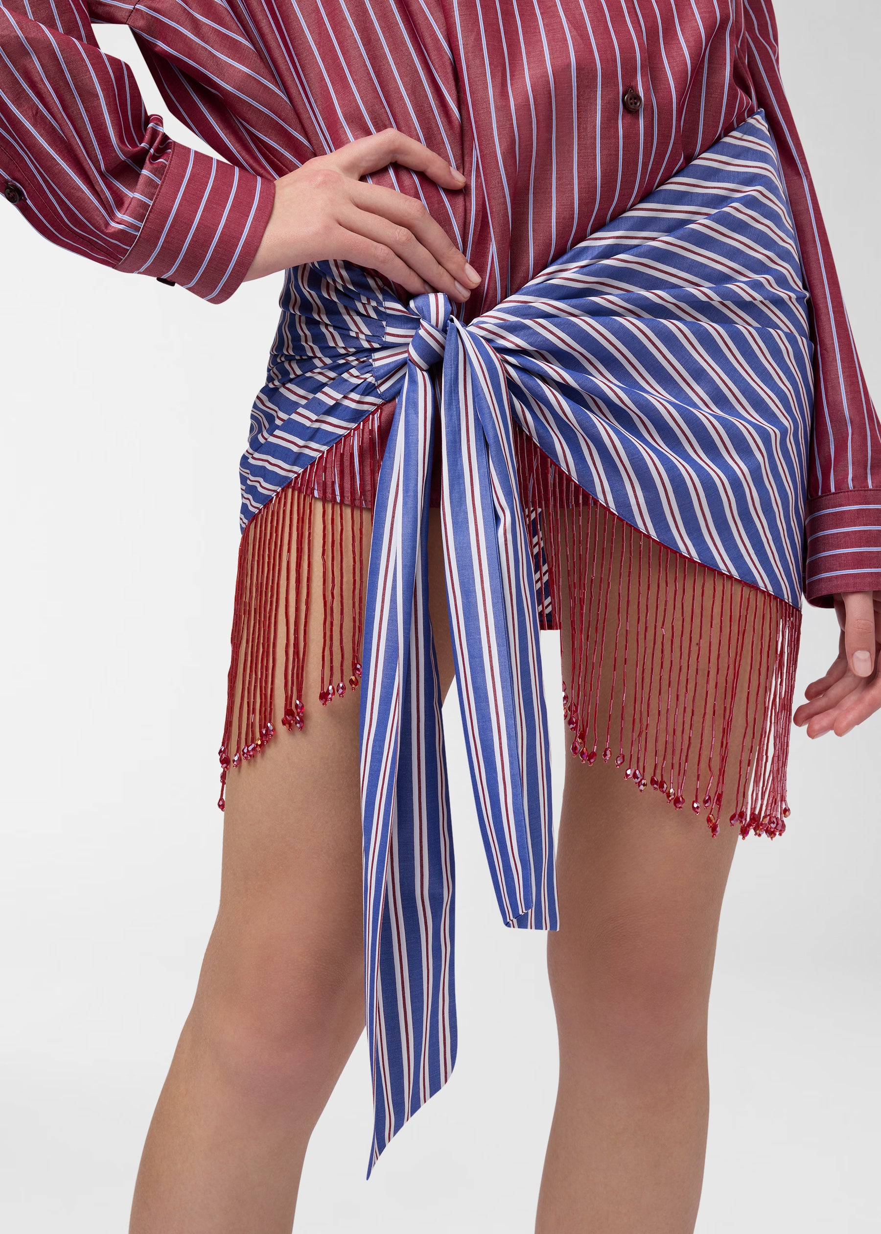 Frontal view — Jolene Mini Dress: model wears rouge noir and light-blue striped shirt dress tied into a knotted skirt at waist, revealing red beaded fringe.