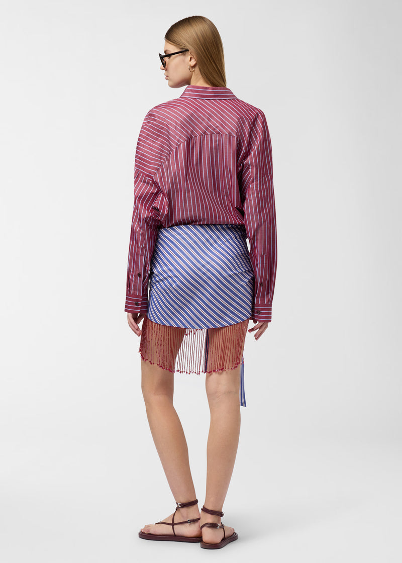Rear view of model wearing Jolene Mini Dress, showing rouge noir and light-blue striped patchwork, pointed collar, contrasting waist detail, and fringed hem with subtle sparkle.