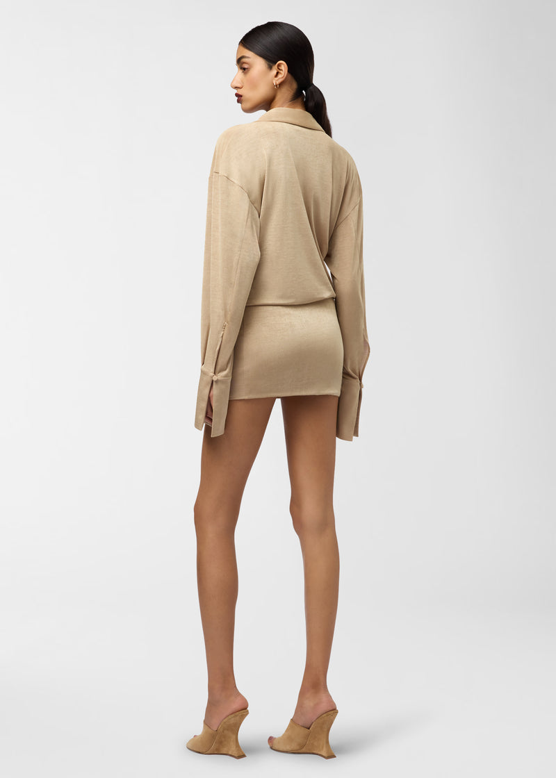 Rear view: Model wearing Lilia Mini Dress stands with head turned, showcasing draped golden jersey, pointed collar and long cuffed sleeves against plain studio background.