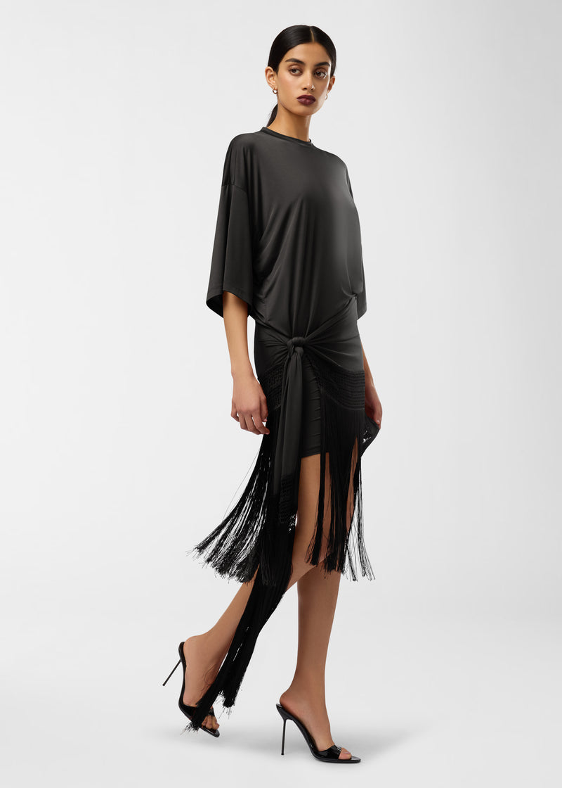 Frontal view: Model wears Azul Midi Dress in black, standing three-quarter pose, loose crew-neck top with butterfly sleeves, side knot cinches waist, long fringe-trimmed hem swaying.
