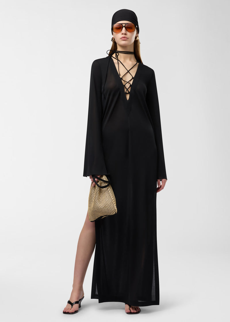 Frontal view — Model wearing Ava Maxi Dress, standing in a studio, showing black semi-sheer jersey-chiffon fabric, lace-up lattice at the chest, flowing kimono sleeves, high side leg slit, holding woven bag.