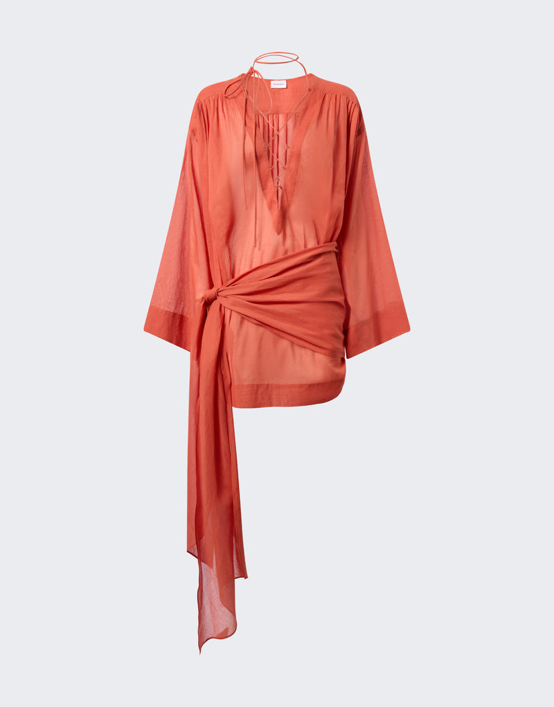 Frontal view — Indira Mini Dress shown hanging on a neutral background, featuring tangerine cotton muslin, long sleeves, stitched pleated shoulders, lace-up chest ties, and a wrap waist panel draping down.