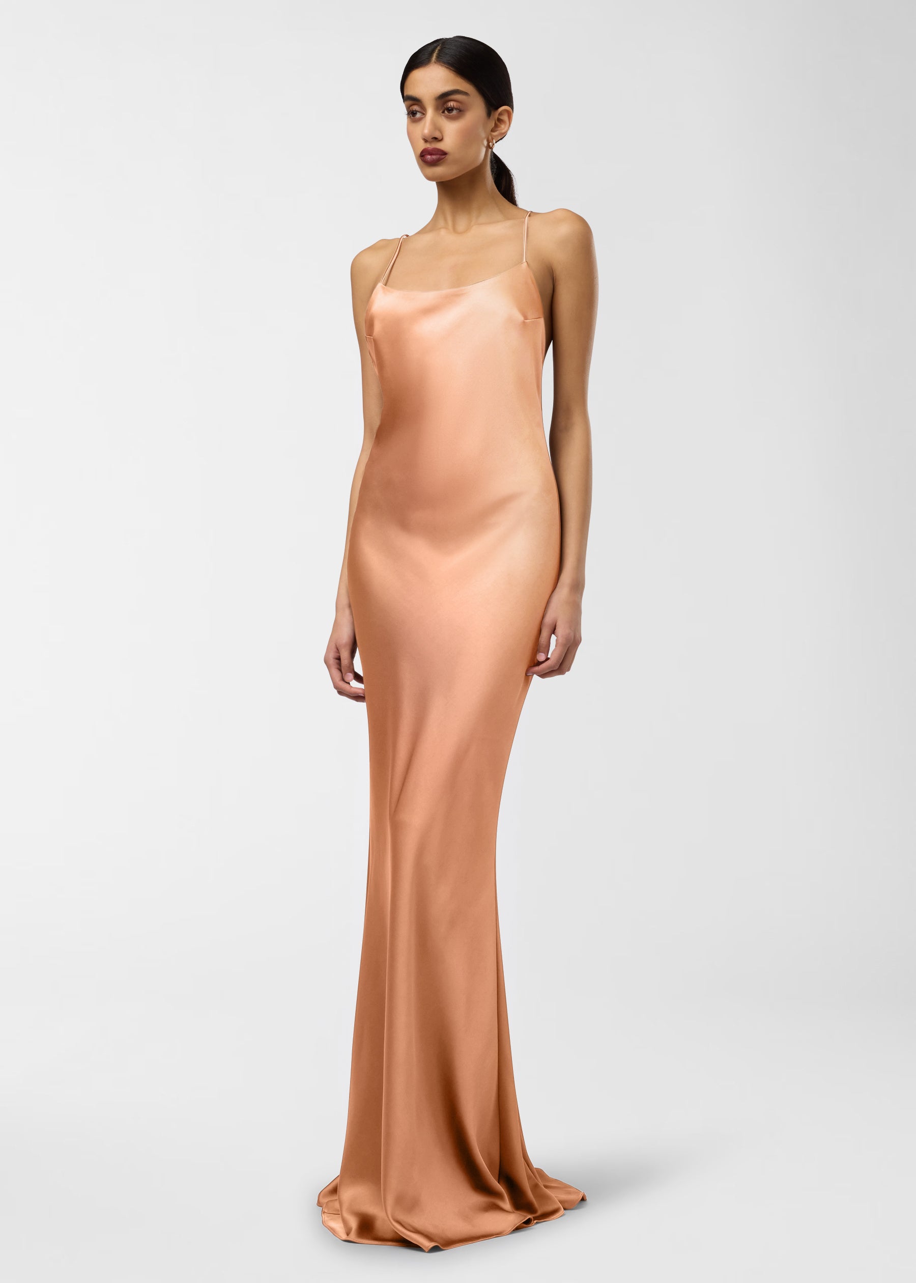 Frontal view — Model wears Ninfea Maxi Dress, standing against a plain background, showing peach liquid satin slip with spaghetti straps, rounded neckline, slim fit and flared mermaid hem.
