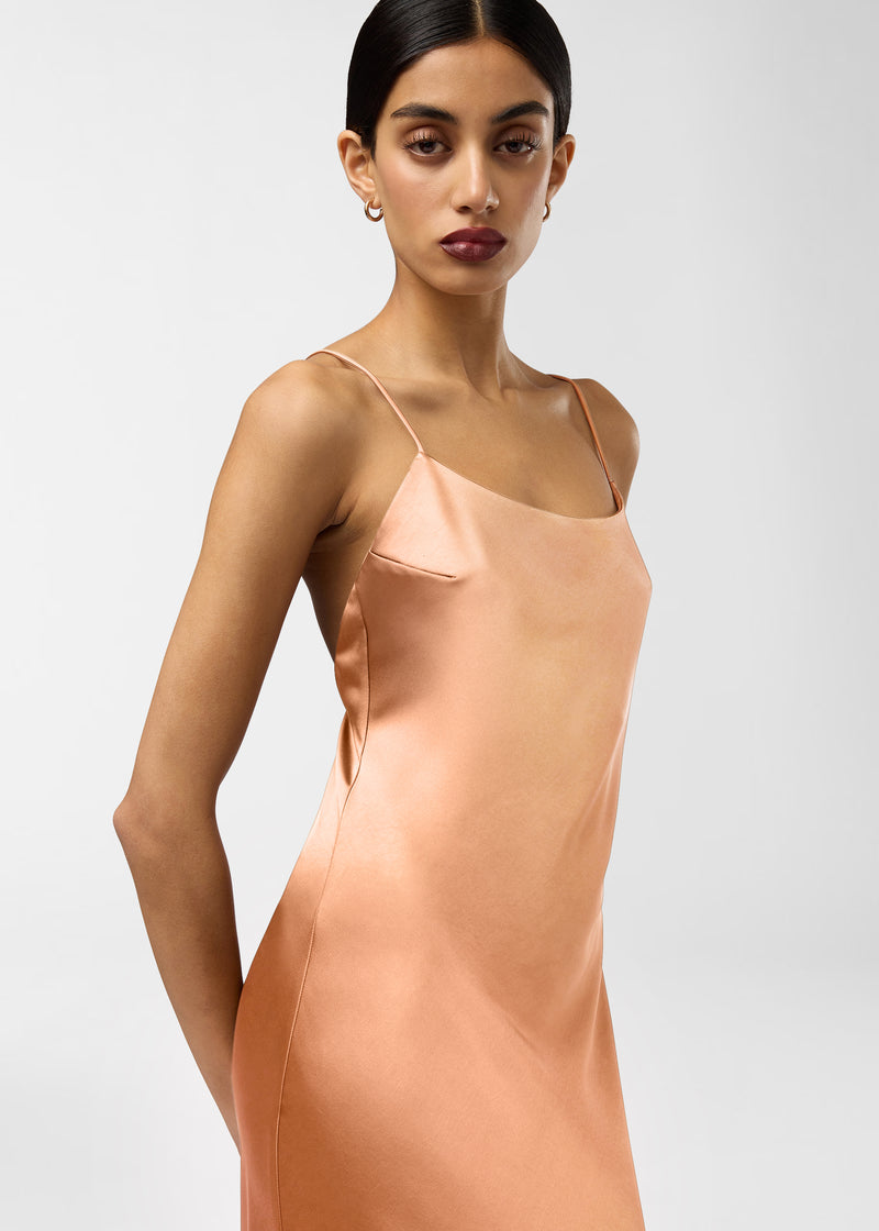 Frontal view: Model wearing Ninfea Maxi Dress standing three-quarter, showcasing peach liquid-satin slip with spaghetti straps, rounded neckline, smooth fitted silhouette and subtle sheen.