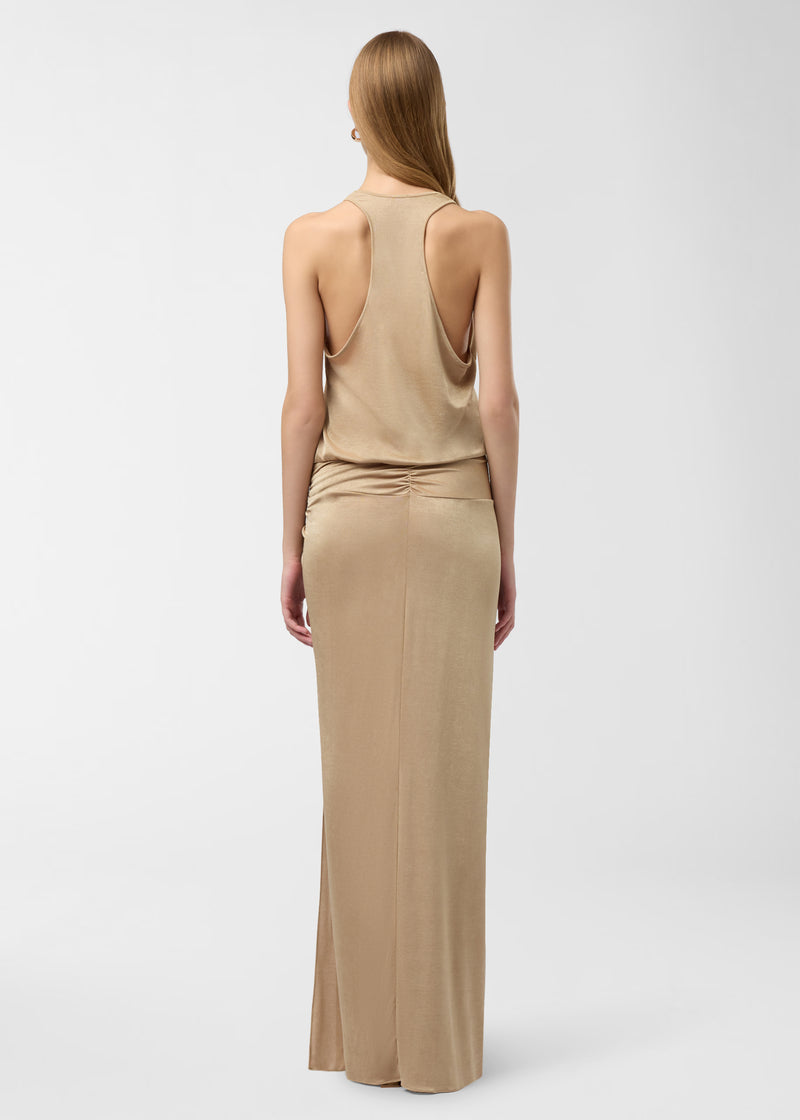 Rear view — model wearing Juno Maxi Dress standing on plain white backdrop, showing plunging racerback, back ruching with gathered hip knot and long golden jersey skirt.