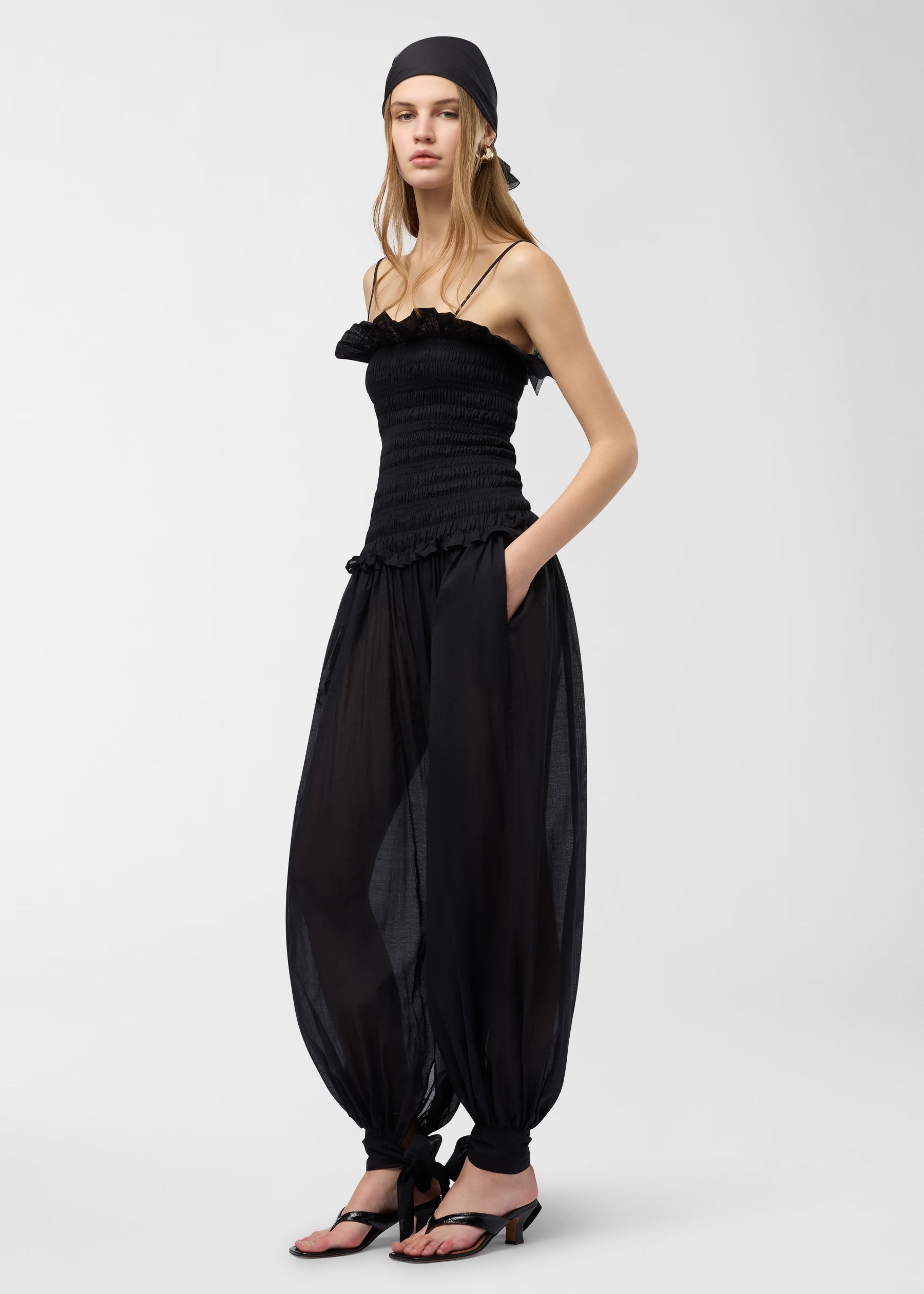 Frontal view — Noor Jumpsuit: model wearing black smocked bodice with frilly chest and waist, spaghetti straps, and semi-sheer balloon pants gathered at tied ankle bows.