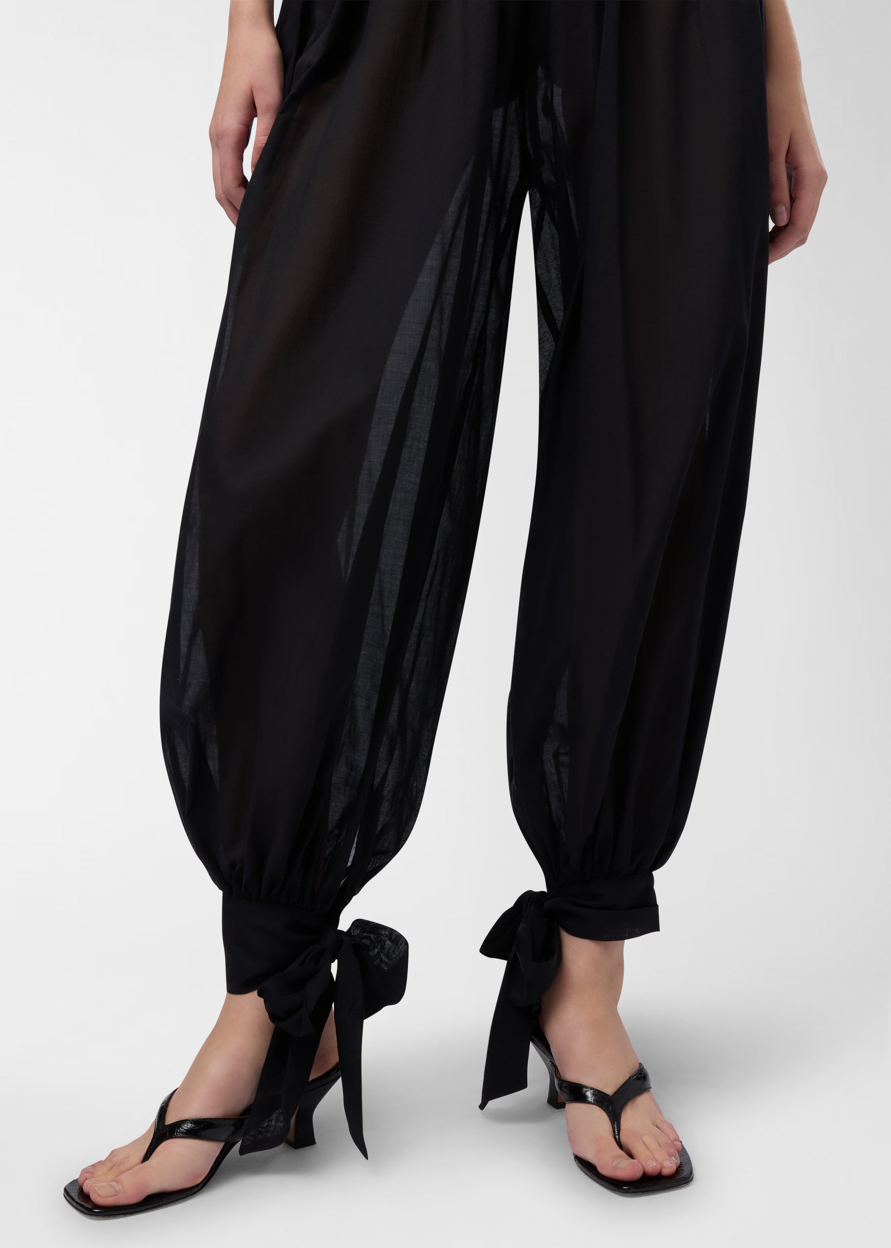 Frontal view of Noor Jumpsuit showing balloon-shaped black muslin pant legs tied in bows at the ankles, semi-sheer fabric revealing leg silhouette and sandals.