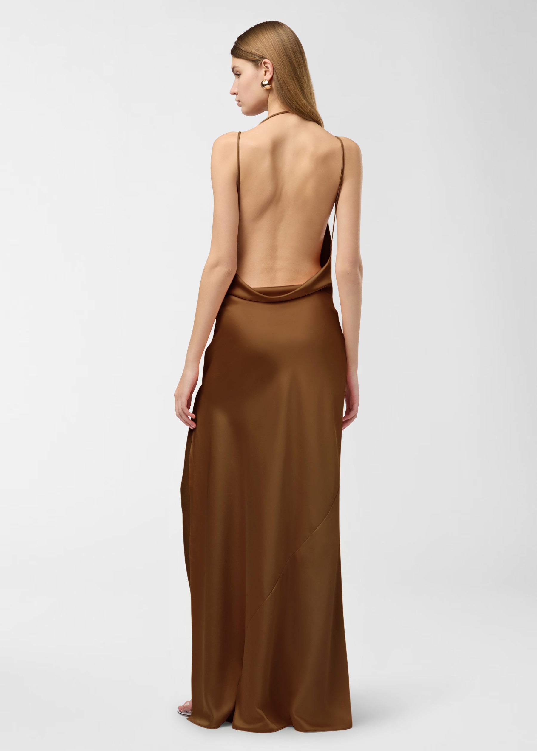 Rear view: Model in Vera Maxi Dress stands, showing open cowl back and flowing bronze satin skirt with high side slit, on a neutral studio backdrop.