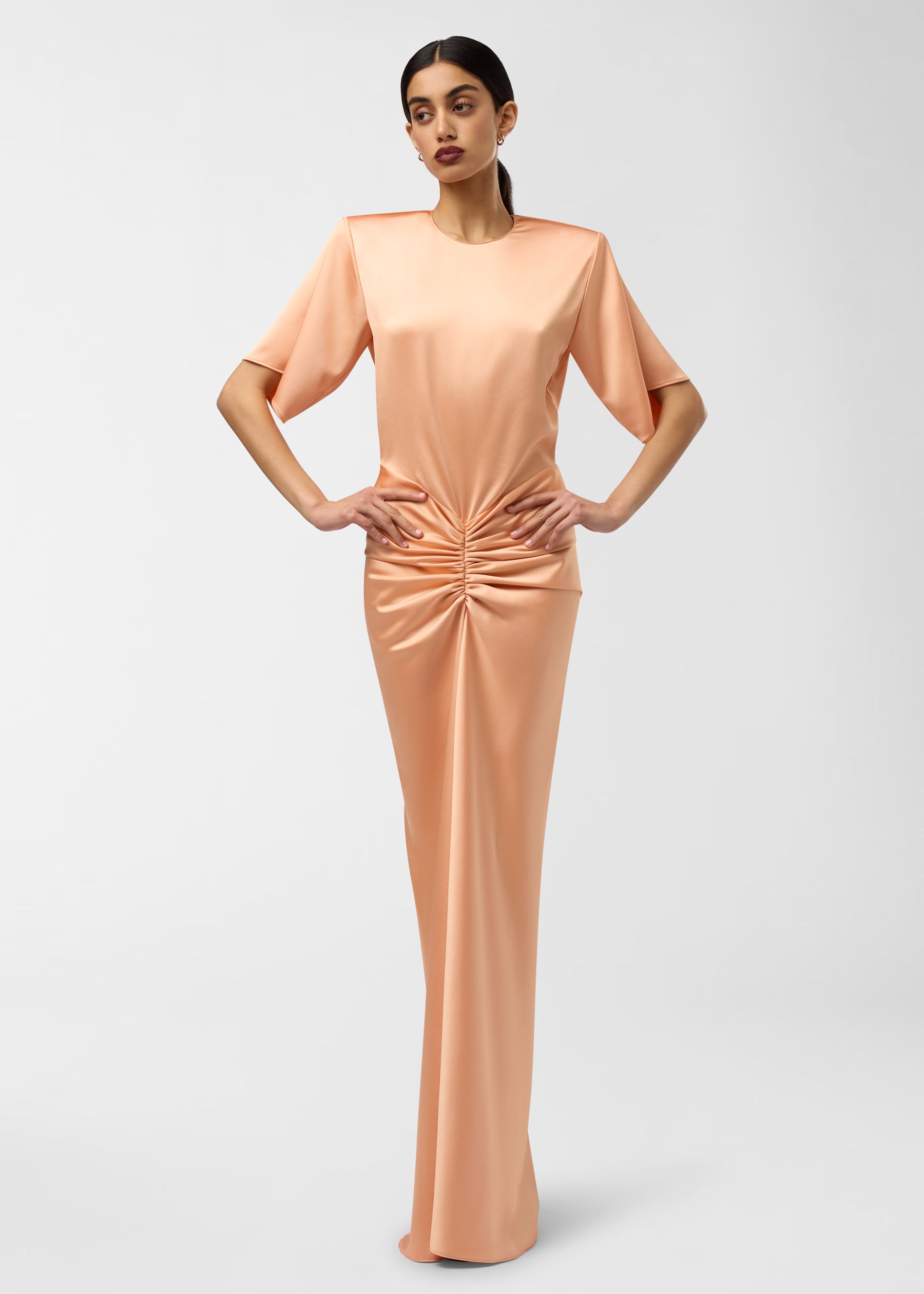 Frontal view — model wearing Iris Maxi Dress in peach: liquid-satin silhouette with structured shoulder pads, elbow-length sleeves and ruched waist, standing hands on hips.