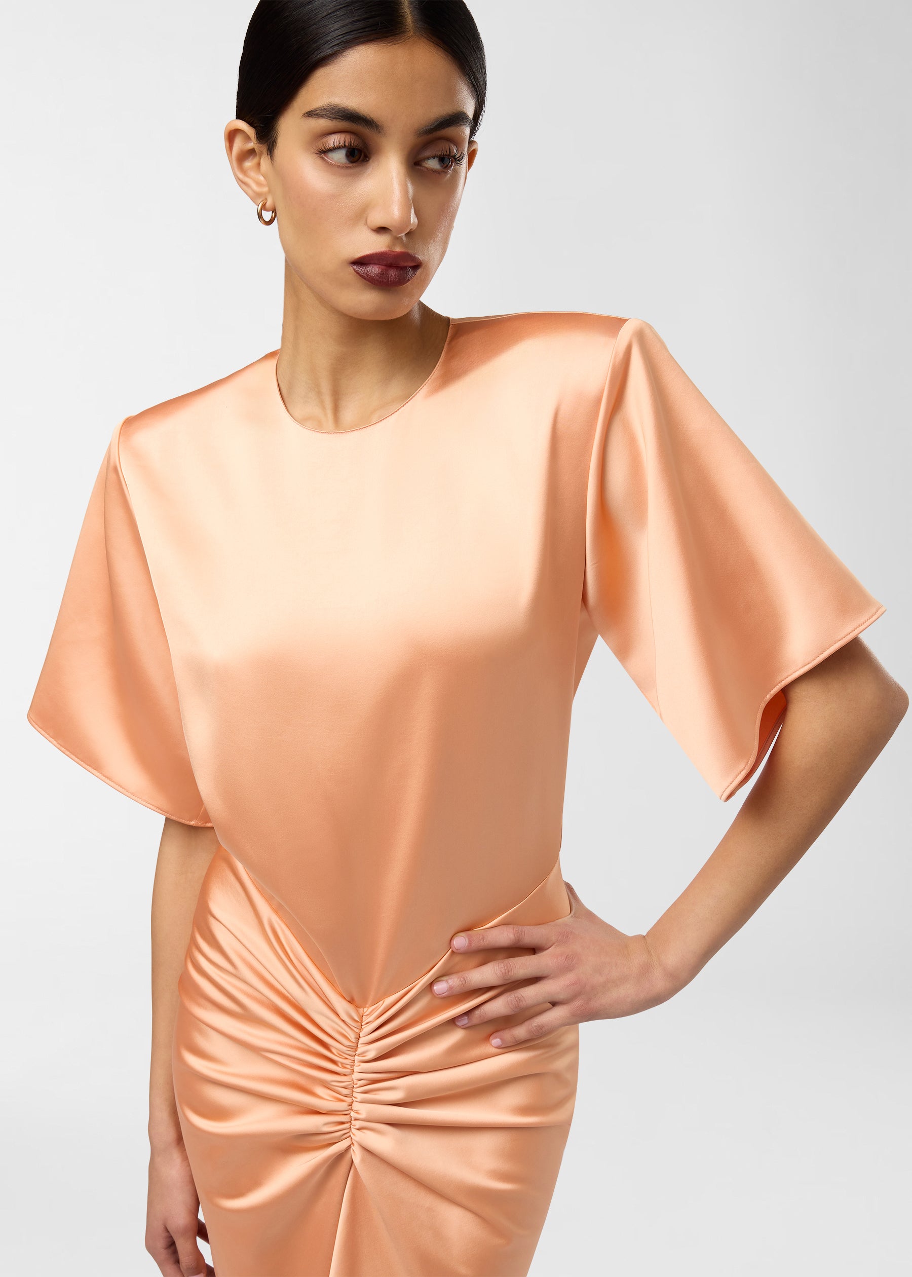 Frontal view — Model wearing Iris Maxi Dress in peach, hand on hip, showing liquid satin sheen, structured shoulder pads, ruched waist and elbow-length sleeves.