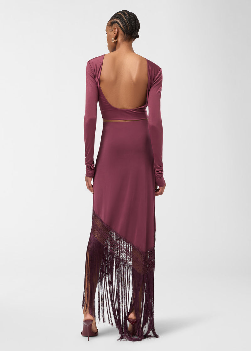 Rear view: Model wearing Marisol Skirt stands, showing high-rise silhouette and asymmetrical hem with openwork weave and long straight fringe in rouge noir against white background.