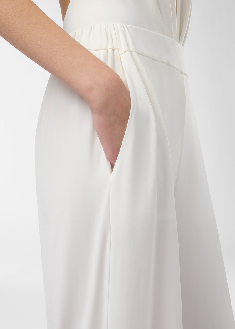 Detail view — Lana Pants show a hand tucked into a concealed side-slit pocket on the high-rise, elasticized waistband; off-white wide-leg fabric drapes to the floor.