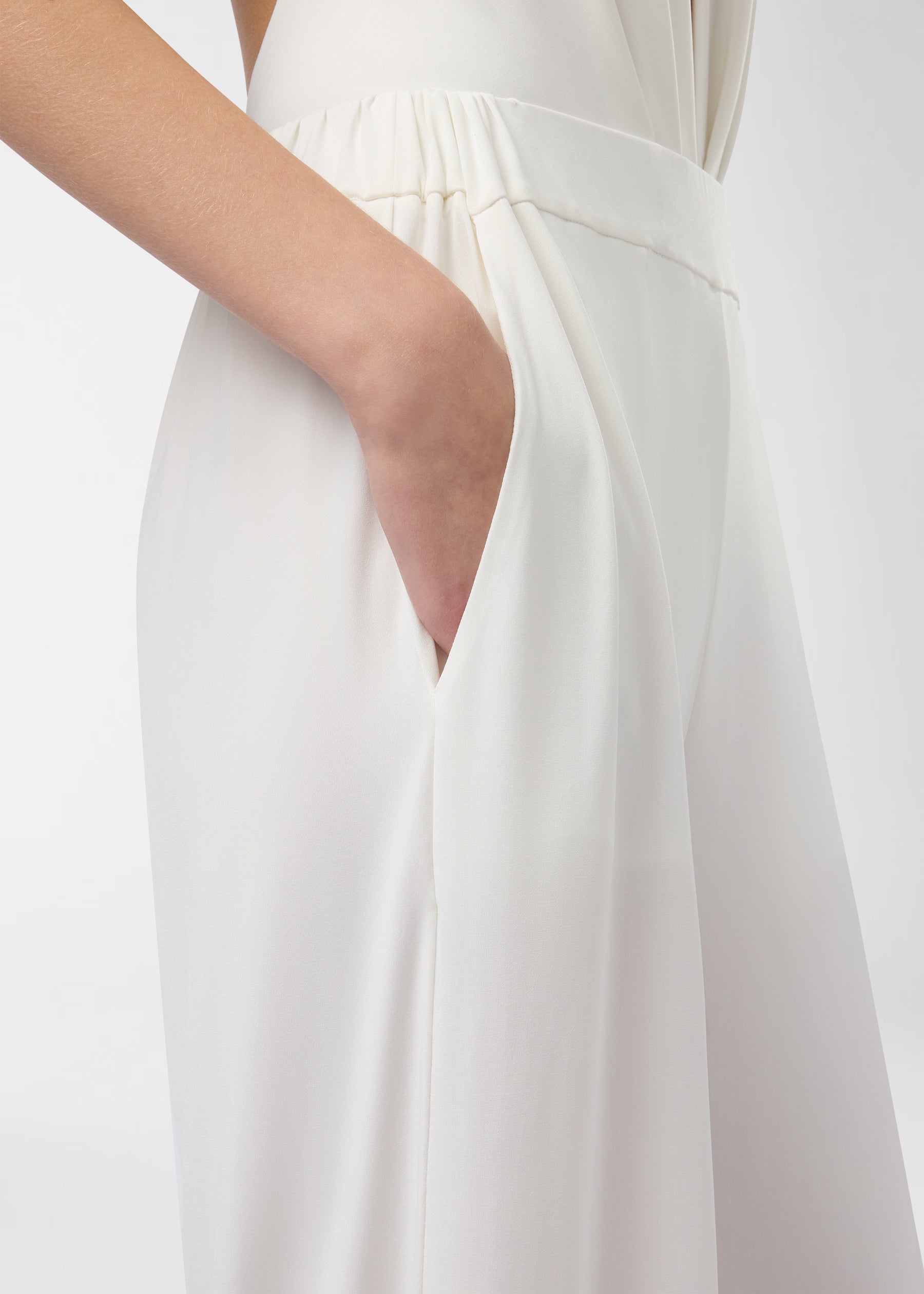 Detail view — Lana Pants show a hand tucked into a concealed side-slit pocket on the high-rise, elasticized waistband; off-white wide-leg fabric drapes to the floor.