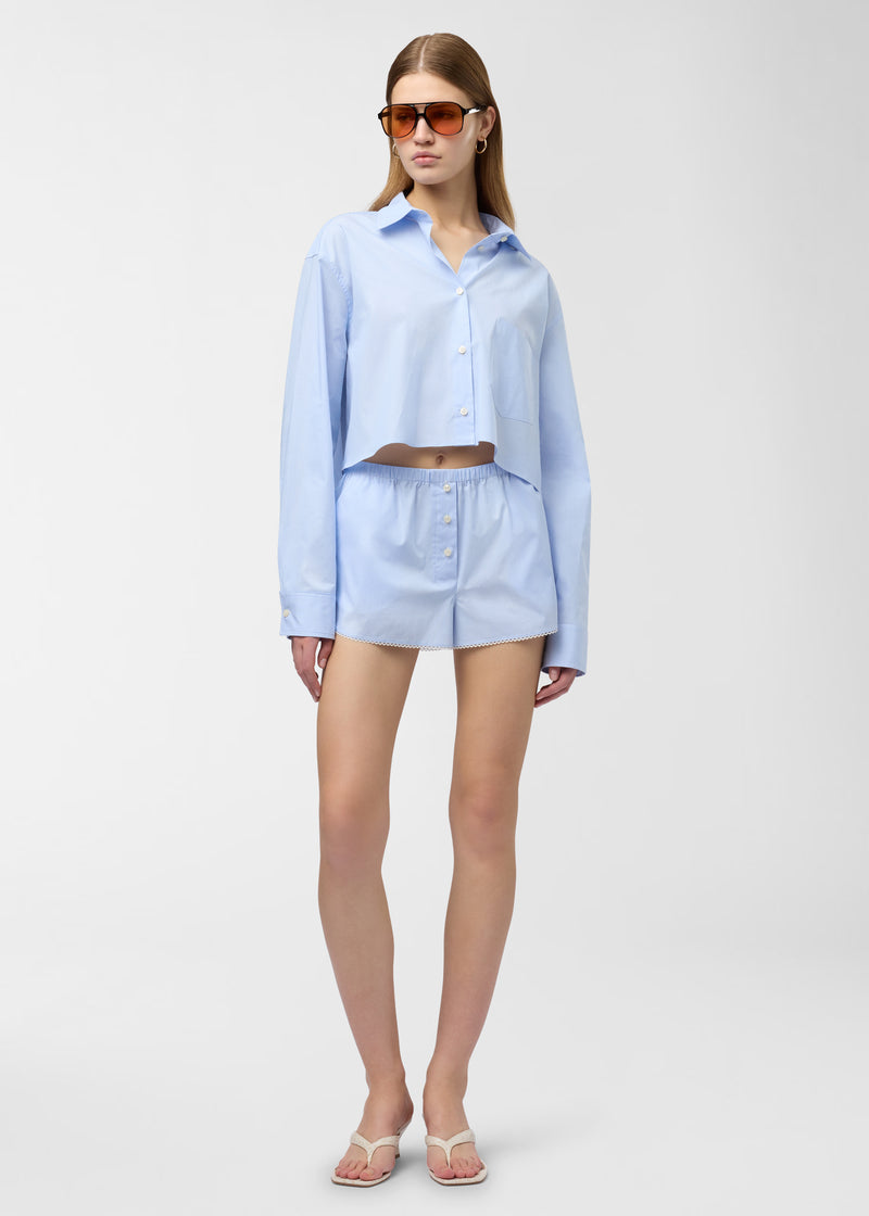 Frontal view: Model wearing Arabella Shorts standing against white backdrop, light blue-and-white micro-striped cotton, elastic low-rise waist, front buttons, micro-lace trimmed hem and side pockets.