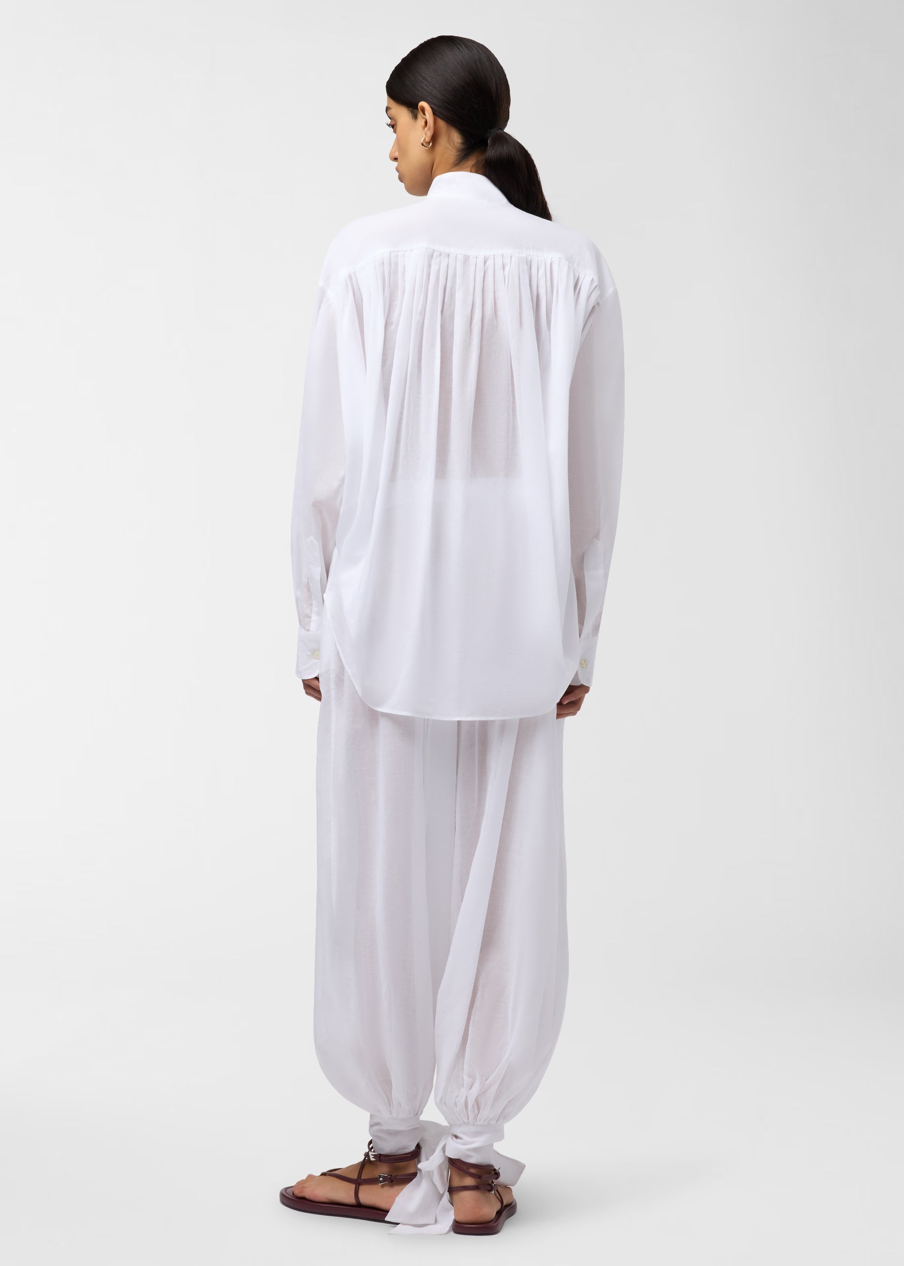 Rear view — Noor Maxi Pants: model wearing white balloon-leg pants tied in ankle bows, paired with oversized white shirt, standing against a plain light-gray background.