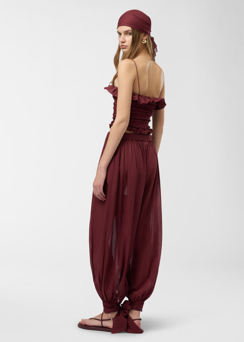 Rear view: Noor Maxi Pants worn by model in rouge noir cotton muslin, wide balloon legs taper and tie into ankle bows, model standing against a plain studio backdrop.