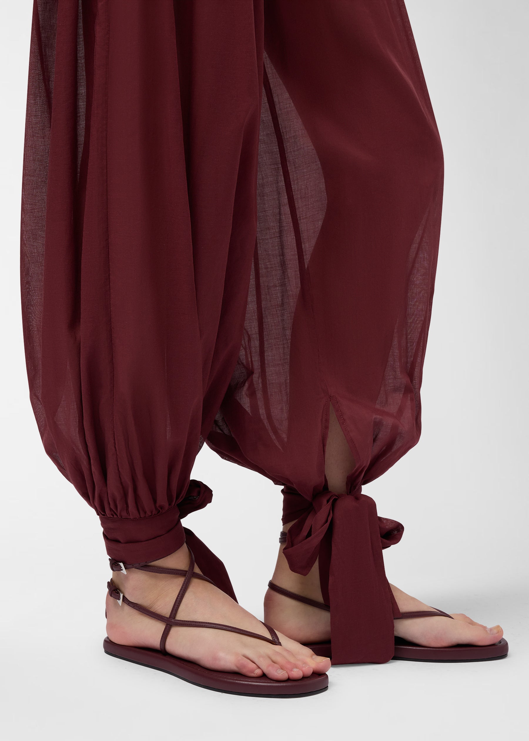 Detail view: Noor Maxi Pants in rouge noir, semi-sheer muslin drapes into wide balloon legs, tapering at ankles tied into bows, paired with strappy sandals.